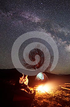 Night camping in mountains. Couple hikers having rest near campfire, tourist tent under starry sky