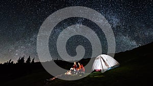 Couple tourists near campfire and tents under night sky full of stars and milky way