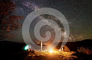 Night camping in mountains. Female hiker resting near campfire, tourist tent under starry sky