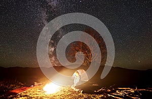 Night camping in mountains. Female hiker resting near campfire, tourist tent under starry sky