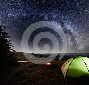 Night camping in the mountains. Illuminated tent and campfire under night sky full of stars and milky way