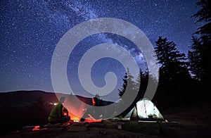 Night camping in mountains near forest with campfire and tourist tent.