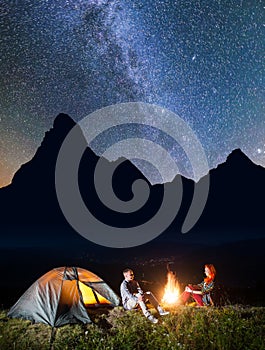 Night camping. Romantic pair sitting near campfire and tent under incredibly beautiful starry sky and Milky way