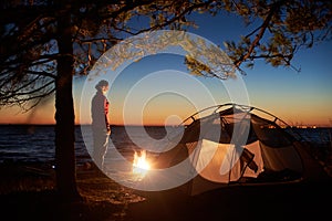 Woman having a rest at night camping near tourist tent, campfire on sea shore under starry sky