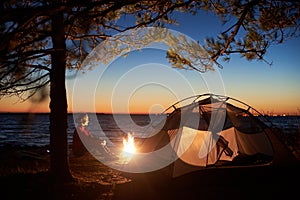 Woman having a rest at night camping near tourist tent, campfire on sea shore under starry sky