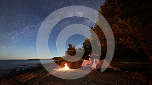 Woman having a rest at night camping near tourist tent, campfire on sea shore under starry sky