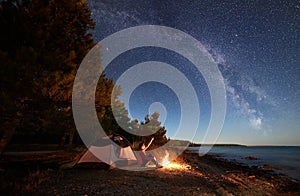 Woman having a rest at night camping near tourist tent, campfire on sea shore under starry sky