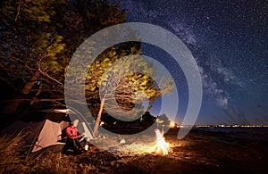 Woman having a rest at night camping near tourist tent, campfire on sea shore under starry sky