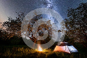 Night camping. tourist tent near campfire under trees and beautiful starry sky and milky way