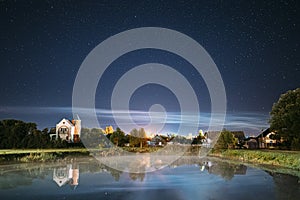 Night Clouds Over River Lake Near Houses In Village. Night Starry Sky Above Lake River With Bright Stars And Meteoric