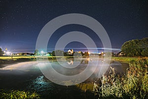 Night Clouds Over River Lake Near Houses In Village. Night Starry Sky Above Lake River With Bright Stars And Meteoric