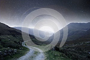 A night concept edit. A mountain track heading into the distance at night with stars in the sky with a bright light glowing on the