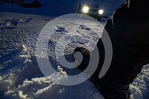 At night. Detail of leg and boot stepping on the snow with many footprints around and an all-terrain car with the lights on,