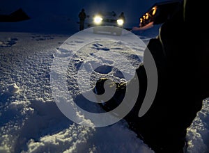 At night. Detail of leg and boot stepping on the snow with many footprints around and an all-terrain car with the lights on,