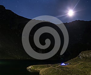 Night mountain landscape with illuminated blue tent. Mountain peaks and the moon. outdoor at Lacul Balea Lake, Transfagarasan,