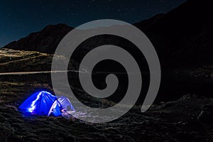 Night mountain landscape with illuminated blue tent. Mountain peaks and the moon. outdoor at Lacul Balea Lake, Transfagarasan,