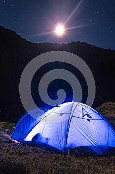 Night mountain landscape with illuminated blue tent. Mountain peaks and the moon. outdoor at Lacul Balea Lake, Transfagarasan,