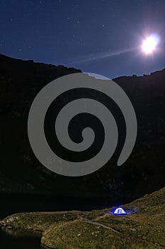 Night mountain landscape with illuminated blue tent. Mountain peaks and the moon. outdoor at Lacul Balea Lake, Transfagarasan,