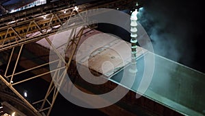 Night panorama of loading grain crops on bulk freighter ship via trunk to open cargo holds at silo terminal in seaport