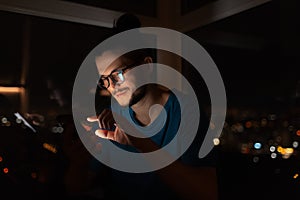 Night portrait of young man touching screen of smartphone. Window background