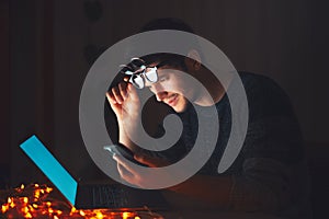 Night portrait of young man wearing round glasses using laptop and smartphone in dark room at home.