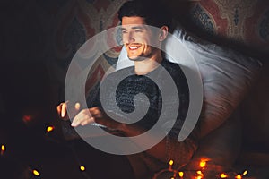 Night portrait of young smiling man lying on pillow on bed with laptop in dark room at home with garlands.