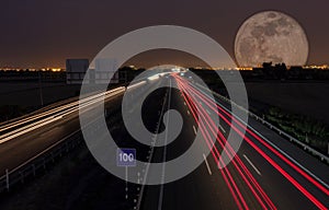 Night road with the city in the background and a spectacular moon. Wakes of the cars