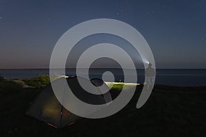 Night scene, camping site, a man with a headlamp stands on a cliff by the sea in the background is a tent