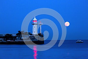 Night seascape with a lighthouse, a large pink moon and a red moon path. The resort city of Yalta in the Crimea on the Black Sea.