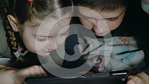 Night shot of teen boy and girl using tablet pc under blanket. Happy children playing with cellphone or smartphone on