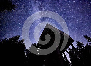 Night sky with the Milky Way over the forest and a bird-watching tower. Trees surrounding the scene