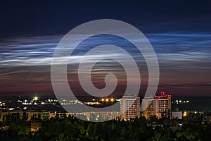 Night sky view of beautiful noctilucent clouds over the city with a cityscape at foreground.