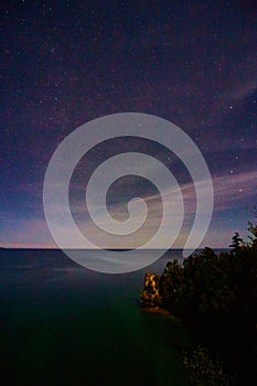 Night sky with whisps of soft pink clouds over Michigan lake and a cliff with a dark forest on the bottom right