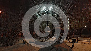 Night snow-covered square with a monument in the form of fish and a street light illuminating the falling snow.