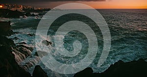 Night stormy ocean by coastal resort town Puerto de Santiago. Sea waves crash on rocky dark volcanic beach at sunset