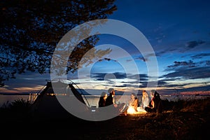 Night summer camping on shore. Group of young tourists around campfire near tent under evening sky