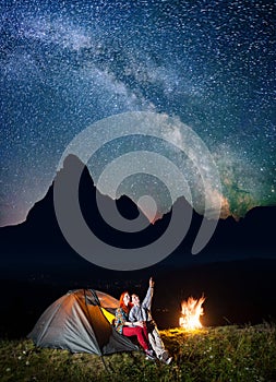 Night tent camping. Happy couple hikers sitting near tent and campfire and enjoying incredibly beautiful starry sky