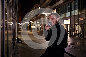 Night time, one young smiling and happy man, 20-29 years, talking over his phone, standing in Autumn coat on street, in business