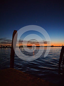 Night on the venice lagoon with mooring posts in silhouette city lights reflected in the sea and illuminated sunset sky