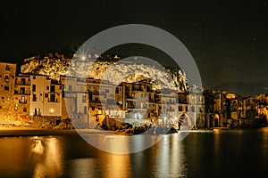 Night view of beach in Cefalu, Sicily, Italy, illuminated old town with colorful waterfront houses, sea and La Rocca cliff.