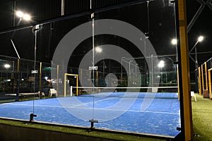 Night view on enclosed blue court for padel with construction created by mesh and the glass back walls.