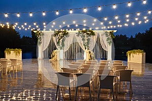 Night wedding ceremony with arch, orchid flowers, chairs and bulb lights in forest outdoors, copy space. Wedding decoration