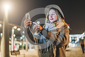 Night. Young woman is standing on city street and is looking at screen of smartphone. Girl uses digital gadget, chatting