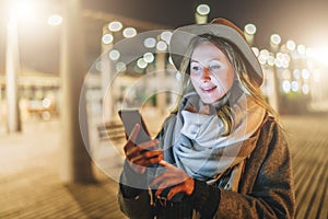 Night. Young woman is standing on city street and is looking at screen of smartphone. Girl uses digital gadget, chatting