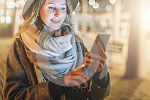 Night. Young woman is standing on city street and is looking at screen of smartphone. Girl uses digital gadget, chatting