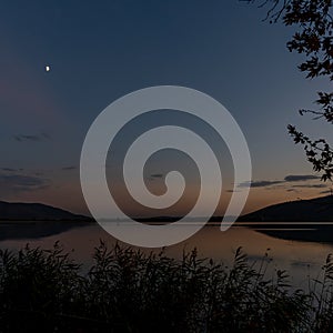 Nightfall over a calm Lake Orestiada in northern Greece with marshgrass in front and the moon in the sky