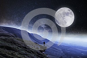 A nightime edit. A hiker on the edge of a cliff surrounded by mountains with the moon and stars above photo