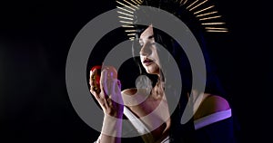 A nightmarish carnival image of a woman in the dark with a crown of shims and a large poisoned red Apple.