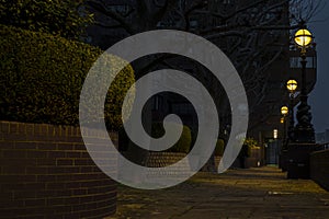 Nighttime on a pathway along red brick wall, hedges and street lamps leaving yellow glow