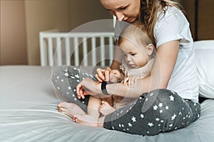 A nine-month-old baby girl with her mother is sitting on the bed and looking at the screen of a smart watch on her mother s hand